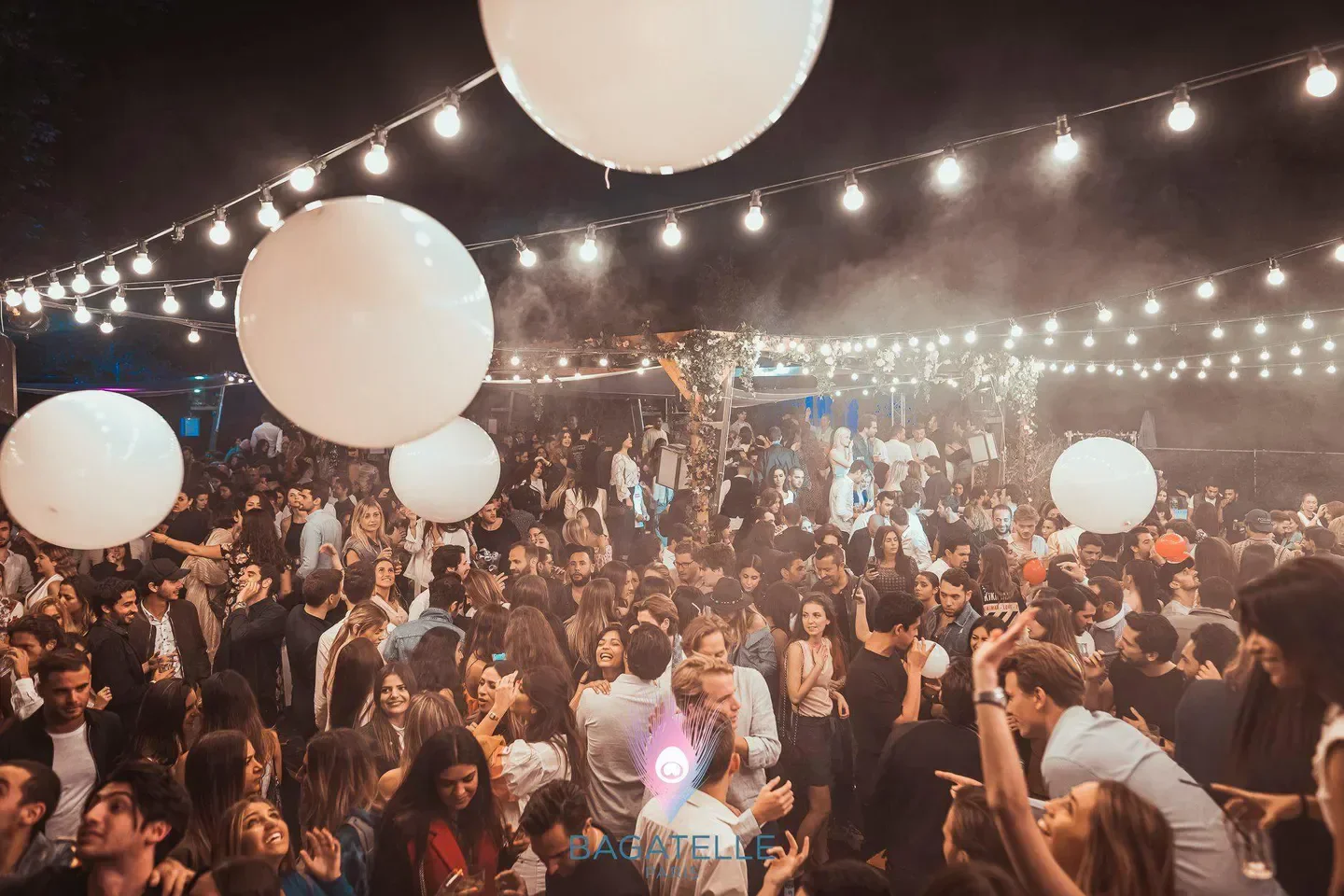 Nightclub scene at Bagatelle Paris, packed with people under string lights and balloons.